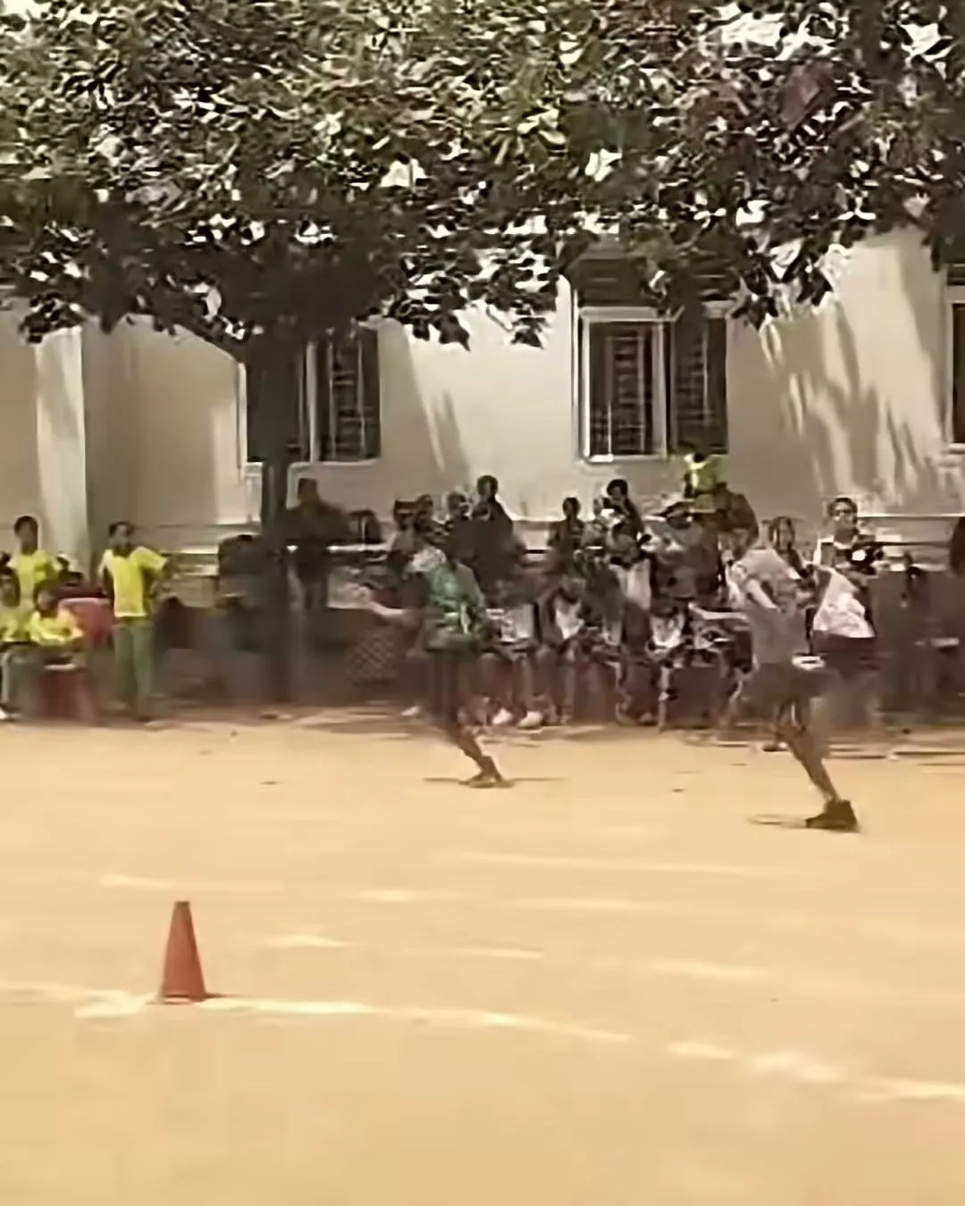 Runners racing along a dirt track as spectators watch from the shade of trees in front of a building