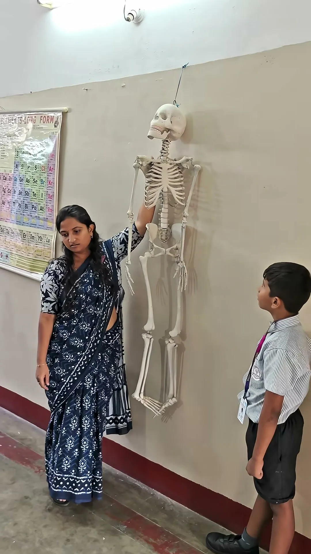 A teacher in a sari holding the arm of a model human skeleton hanging on the wall as a student looks on