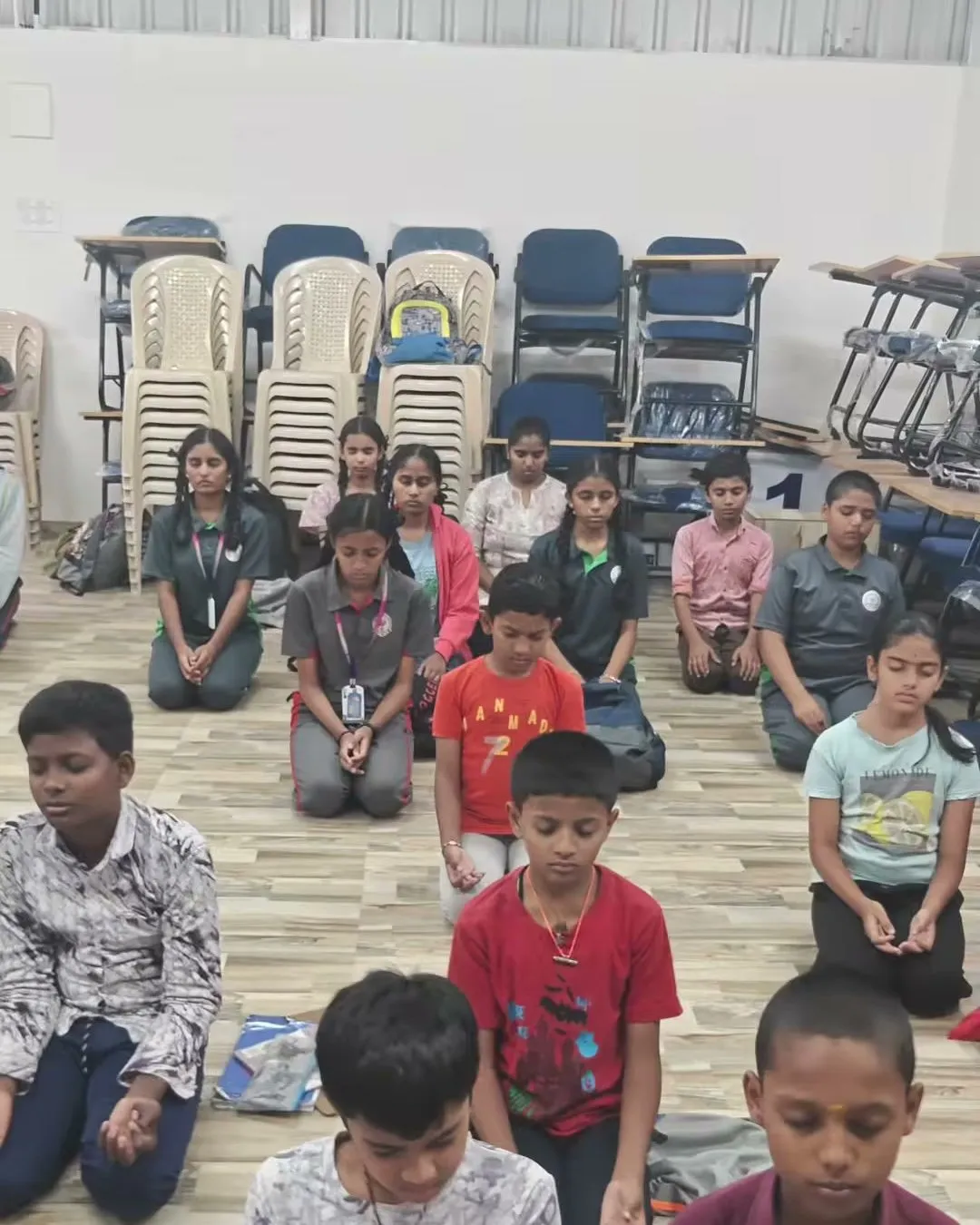 Children kneeling in Vajrasana with their eyes closed on the floor of a hall, with stacked chairs behind