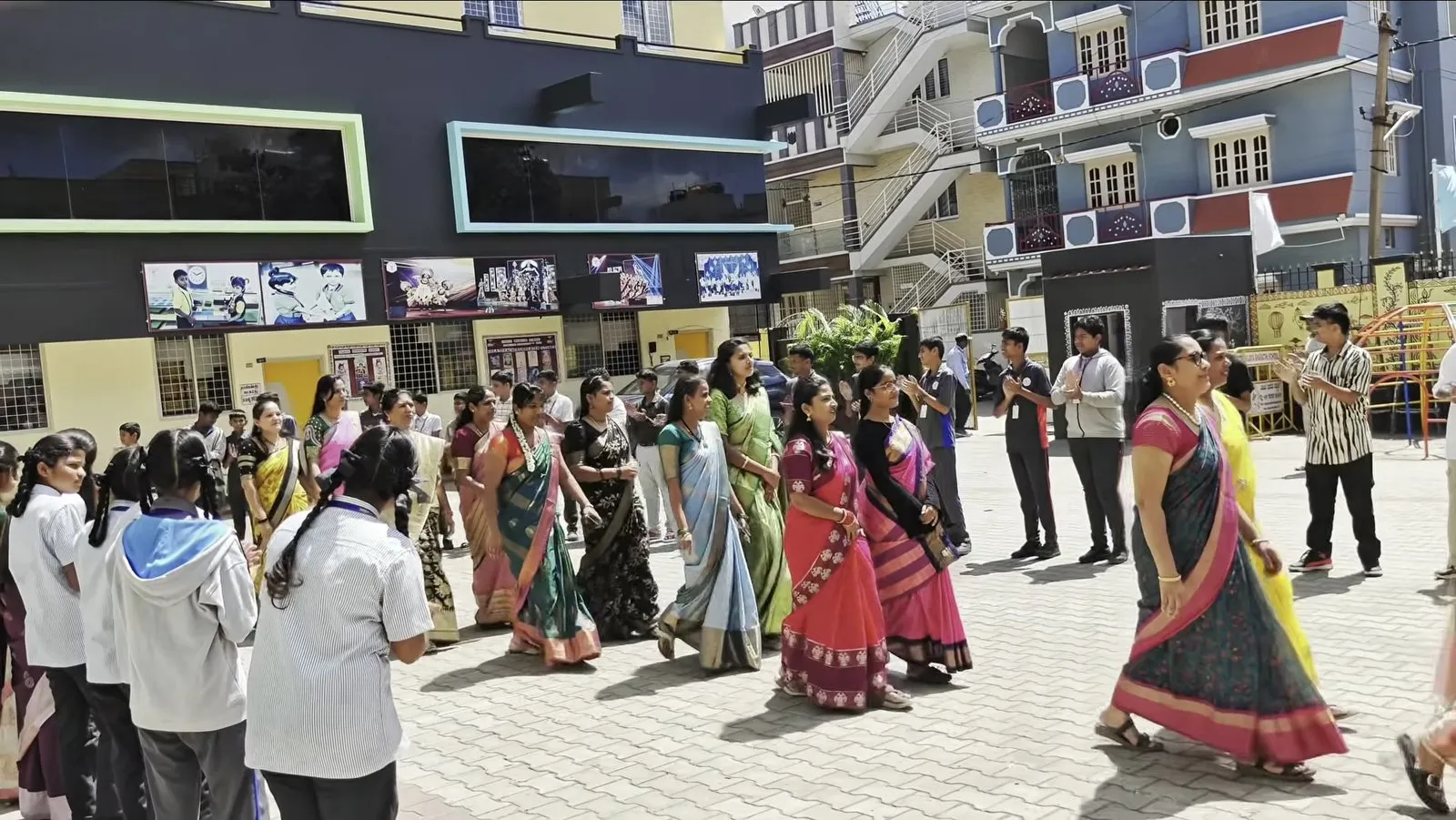 Students in a circle in the school courtyard, with people in saris walking around the inside of the circle