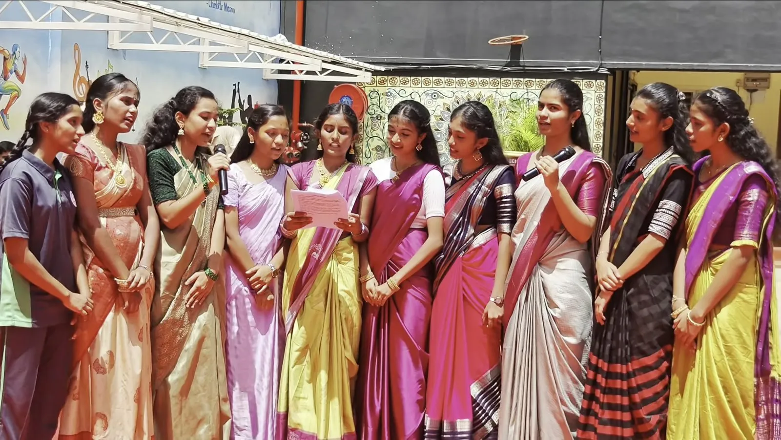 A line of young women in saris in the courtyard, one speaking into a microphone and another reading from a sheet of paper