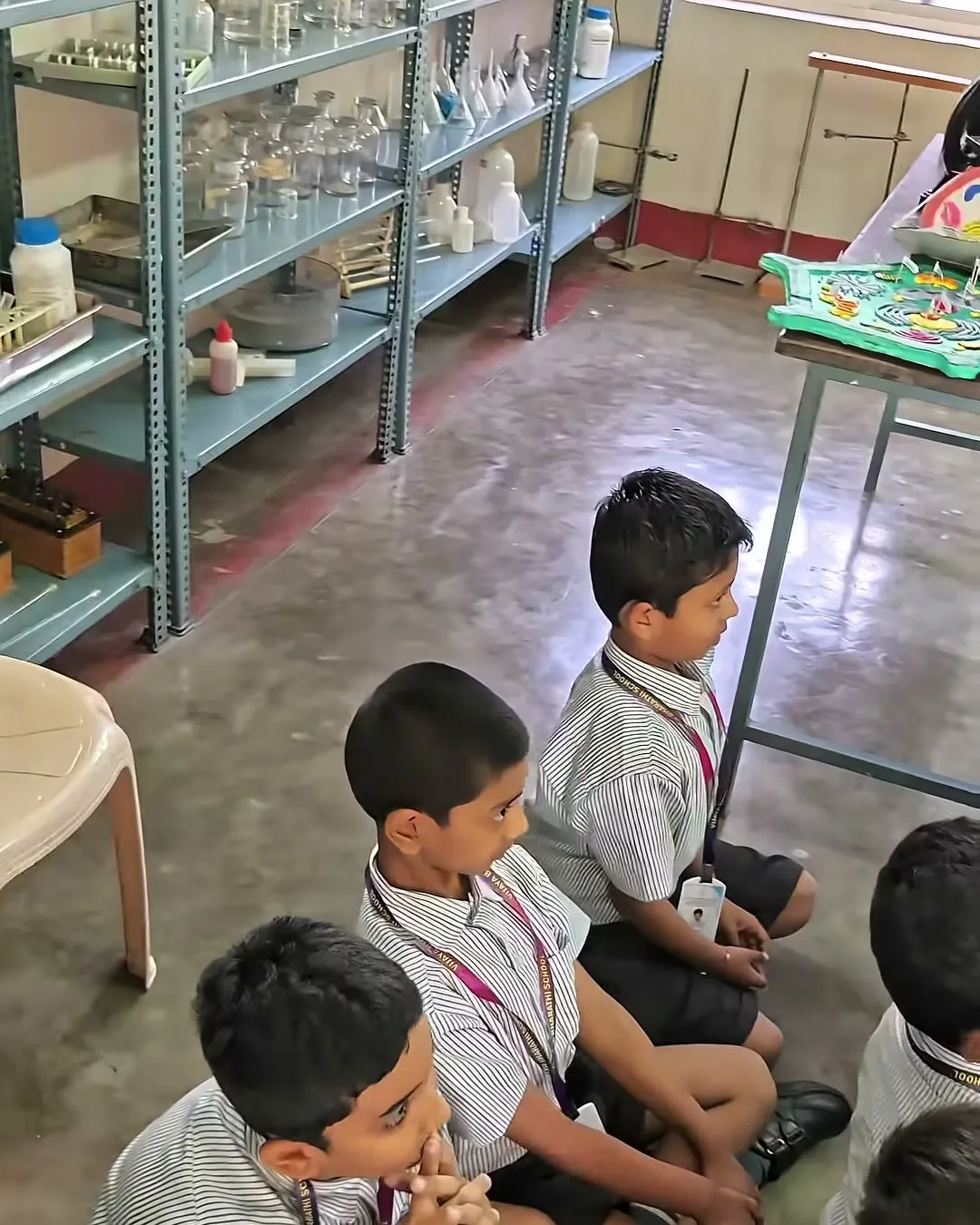 Young students sitting on the floor of a science laboratory, with shelves of glassware and equipment behind