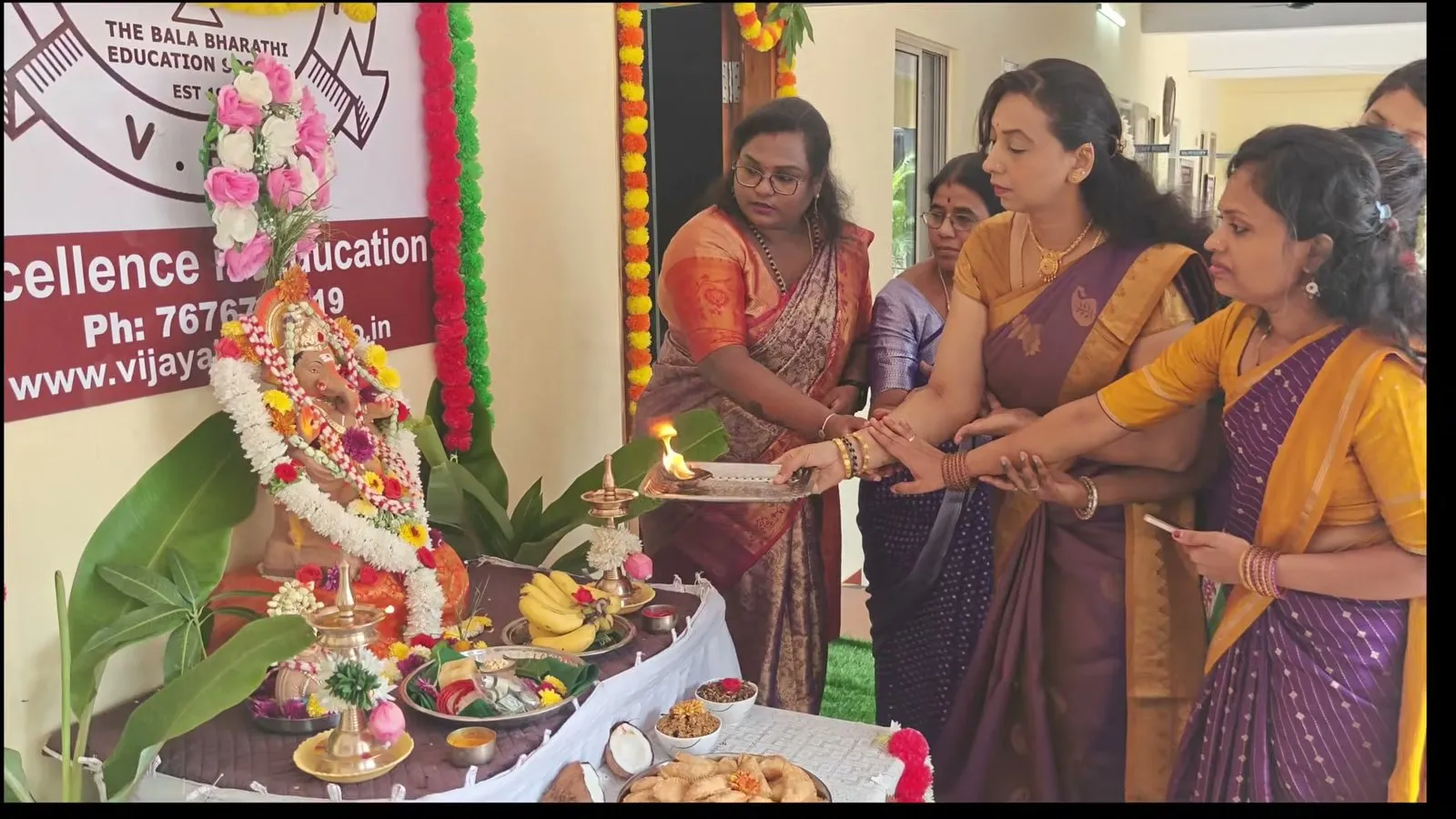 Women in saris holding a lit lamp together in front of the Ganesha idol and plates of fruit and offerings