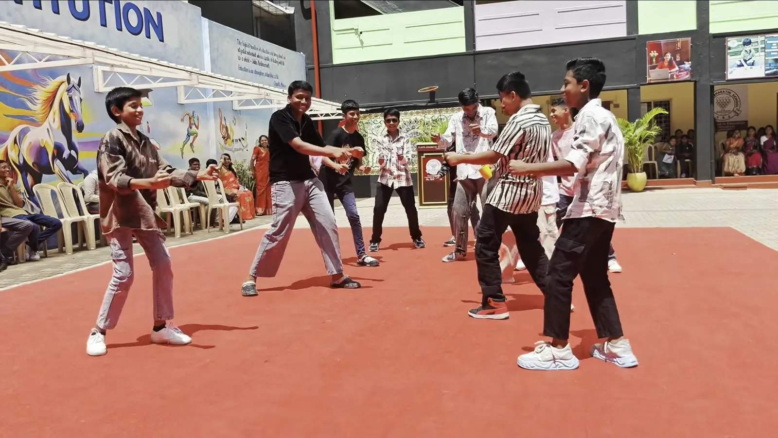 Boys dancing on a red carpet in the courtyard, with painted murals and seated spectators behind