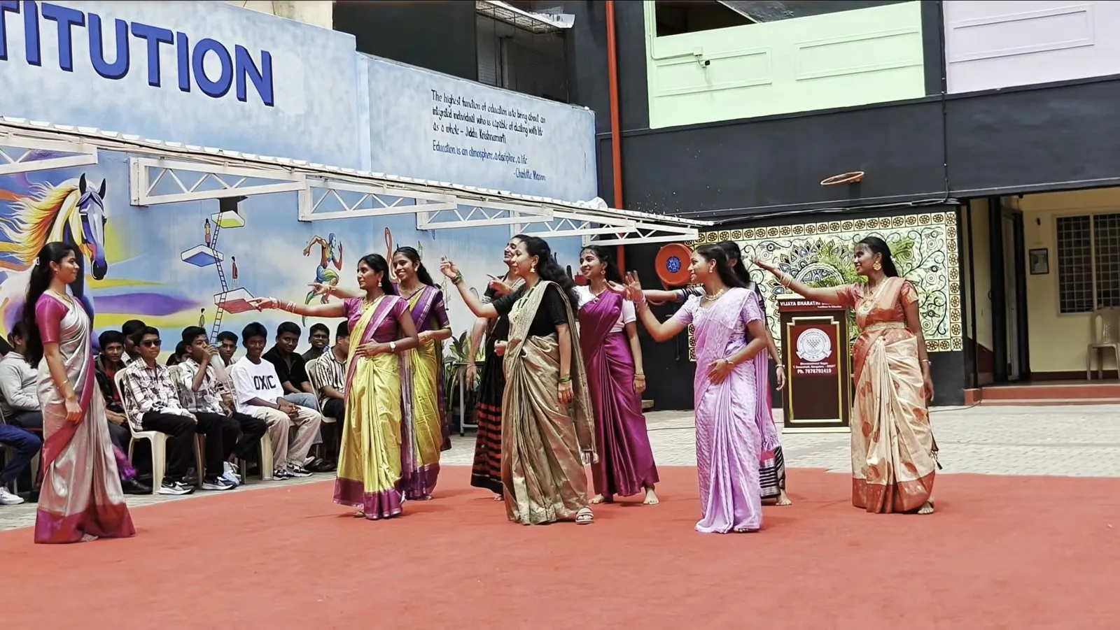A group in saris dancing on a red carpet while students watch from chairs beside a painted mural