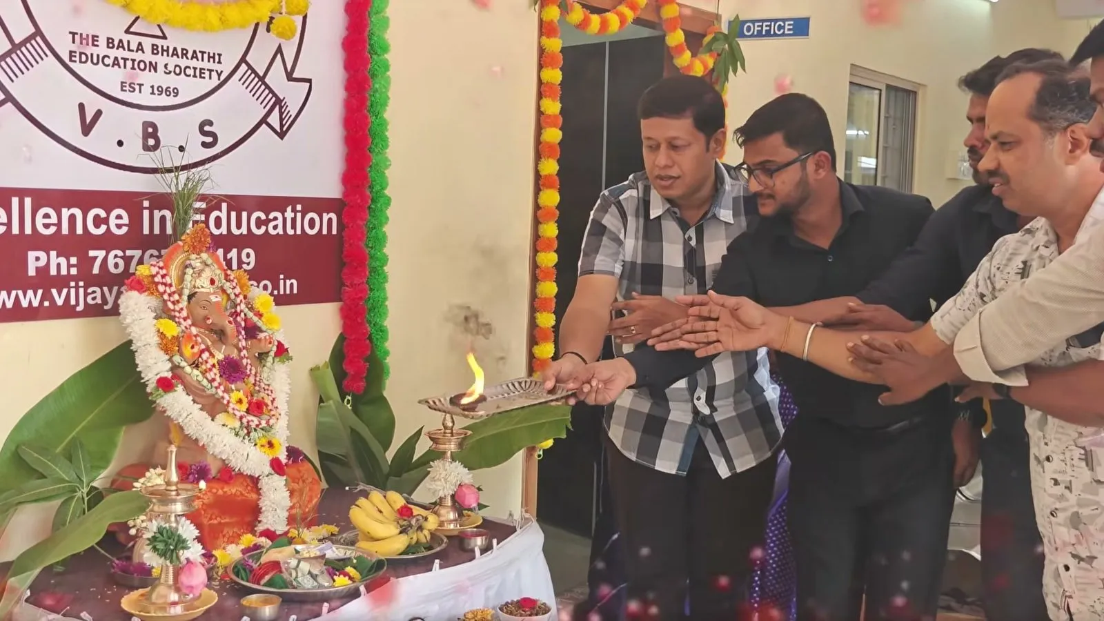 Men holding a lit lamp together in front of the Ganesha idol, with a school crest banner behind