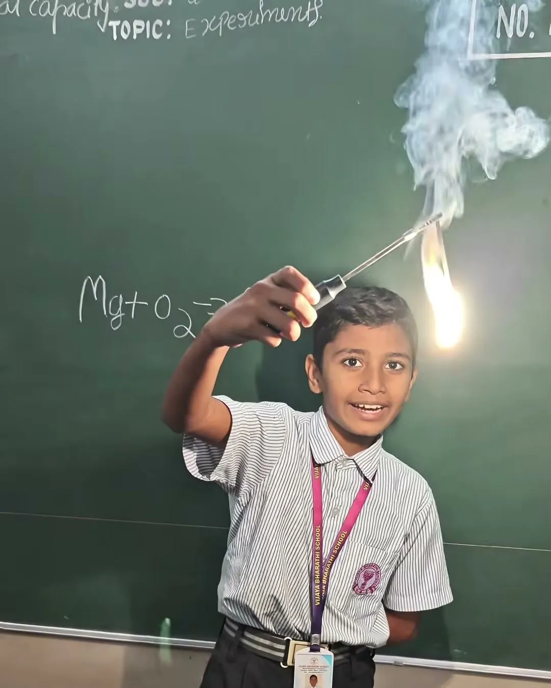 A student holding a burning strip of magnesium ribbon with tongs in front of a blackboard showing the equation