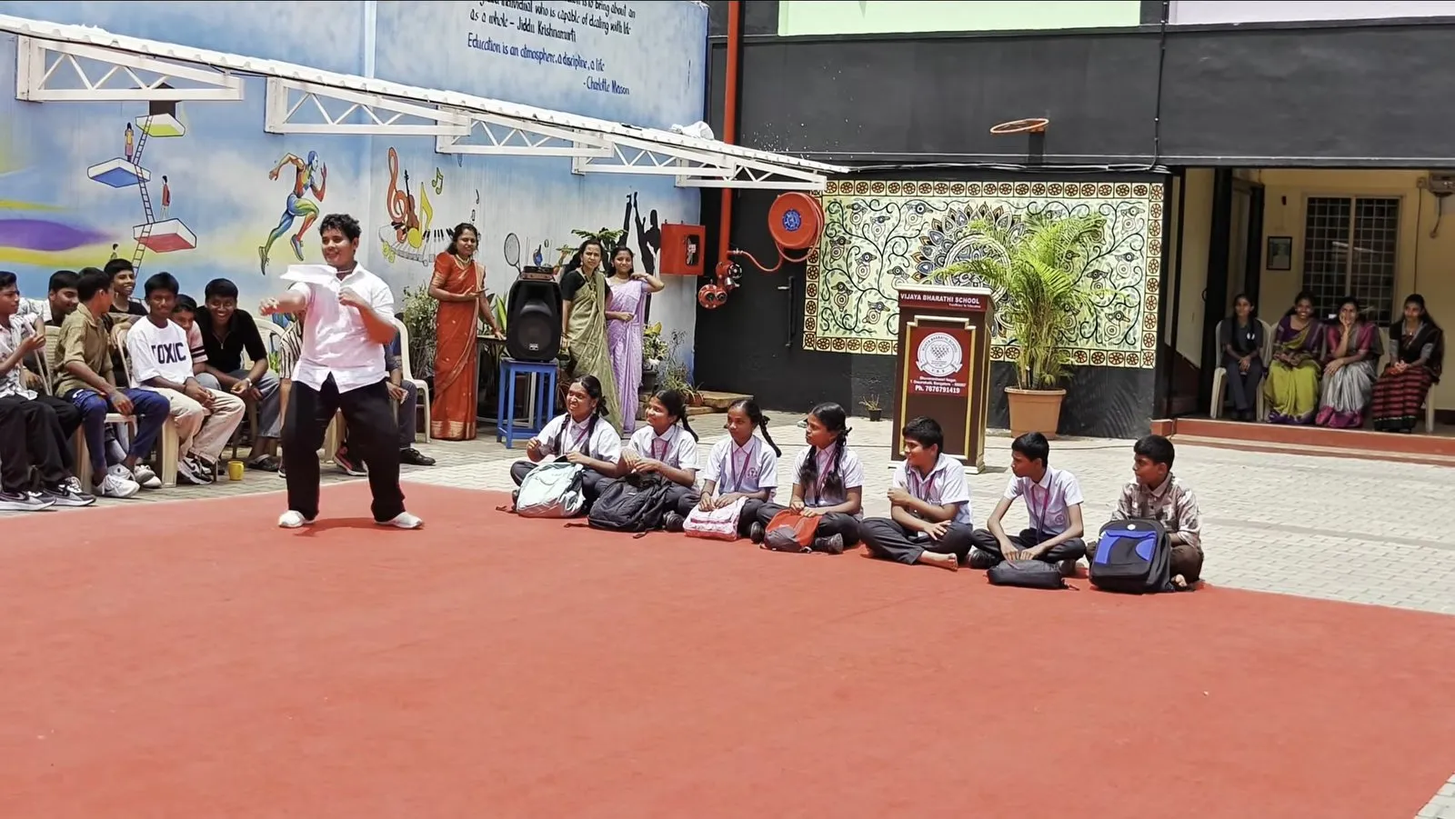 A student performing on a red carpet beside a row of seated students, with painted murals and a lectern behind