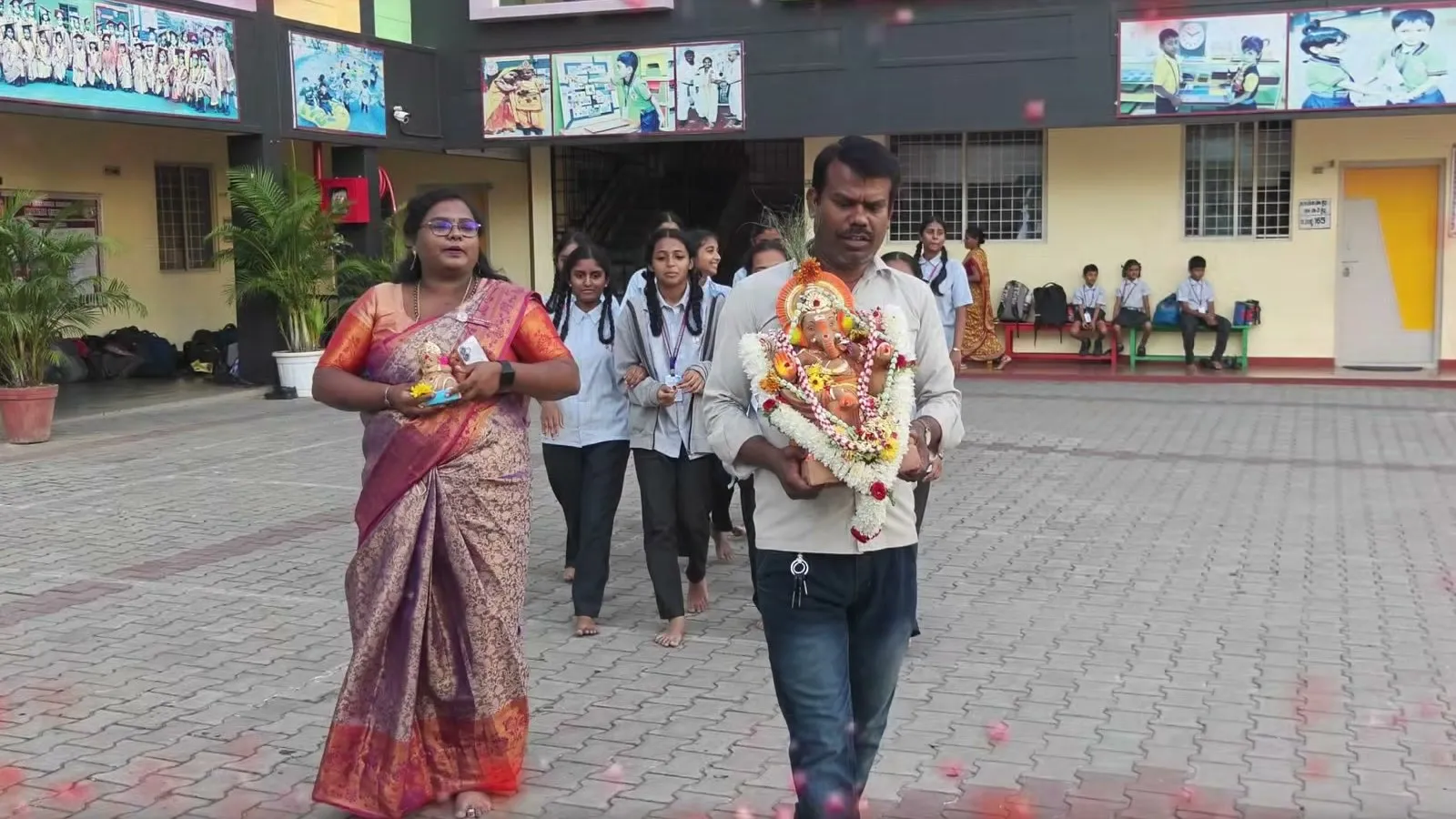 A man carrying the Ganesha idol across a paved courtyard, walking beside a woman in a sari, with students following