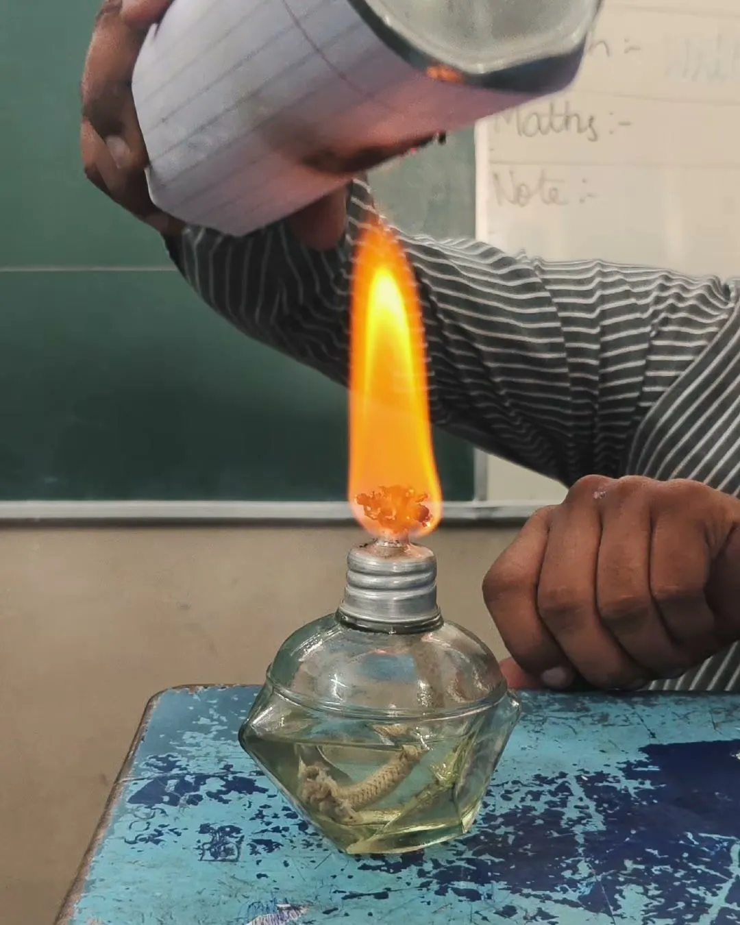 Hands holding a paper-wrapped metal bottle over the flame of a small glass lamp