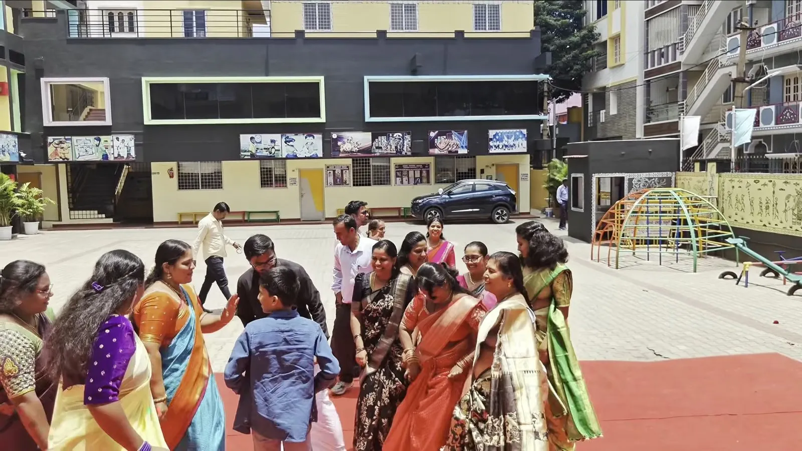Women in saris and men in shirts playing a group game in the courtyard, with school buildings and a play frame behind