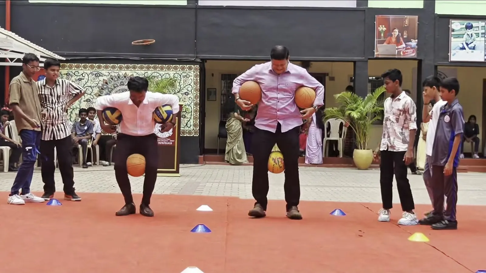 Two men holding balls and balancing another between their knees on a red carpet marked with cones, as students watch