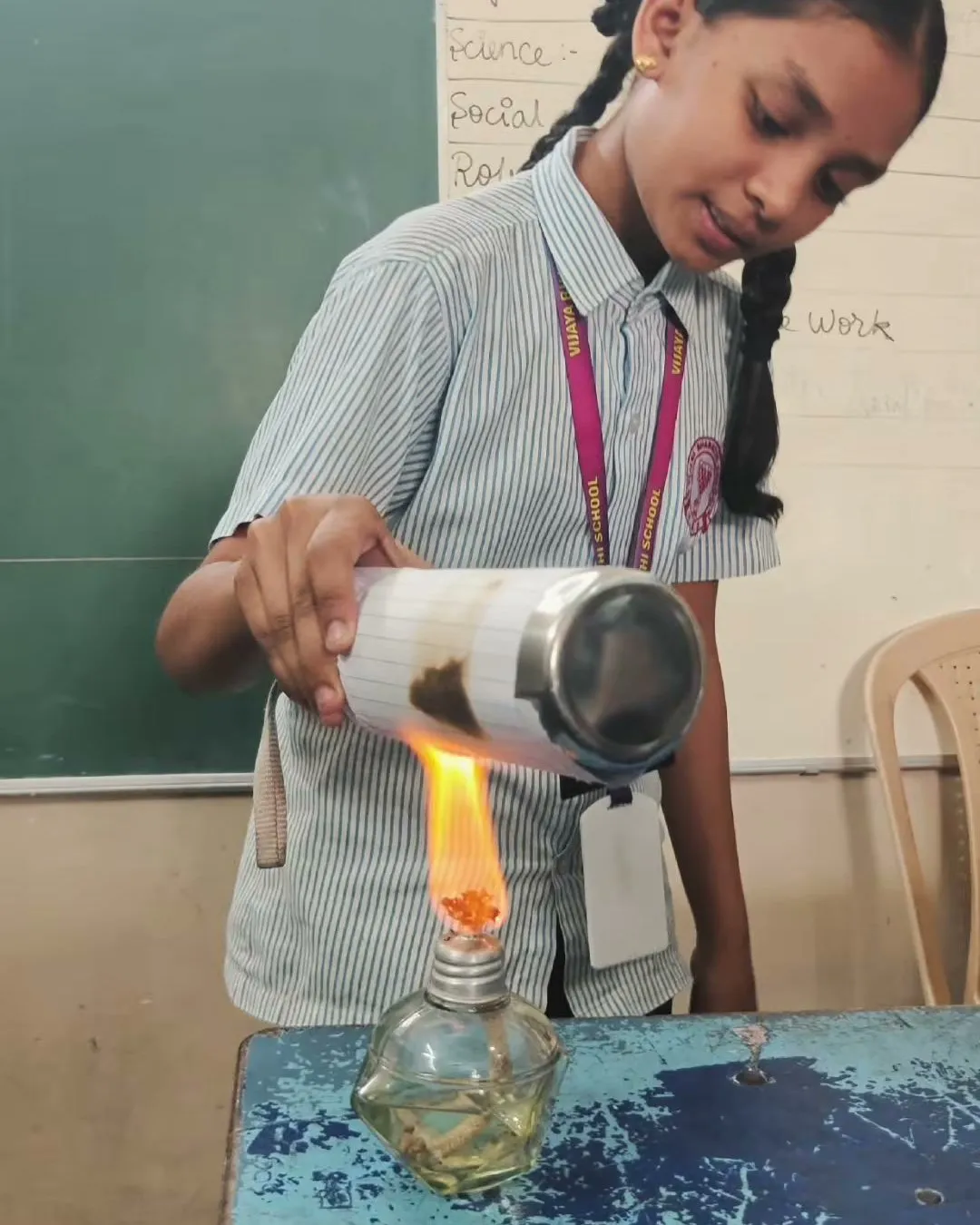 A student holding a paper-wrapped metal bottle over a small lamp flame at a desk