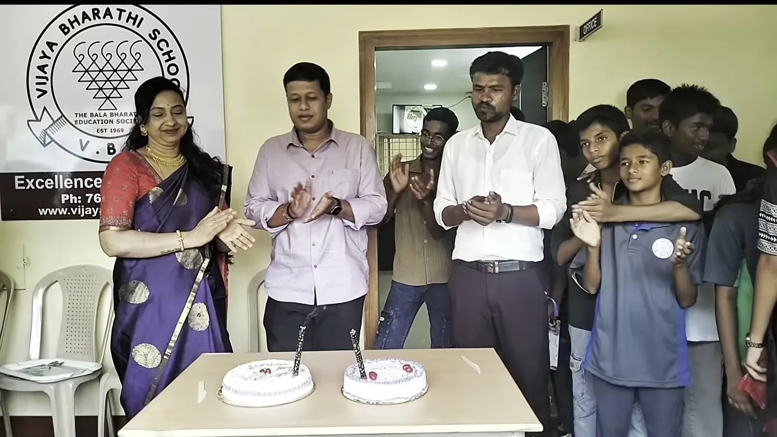 Adults and students standing around a table with two cakes, in front of a banner showing the school crest
